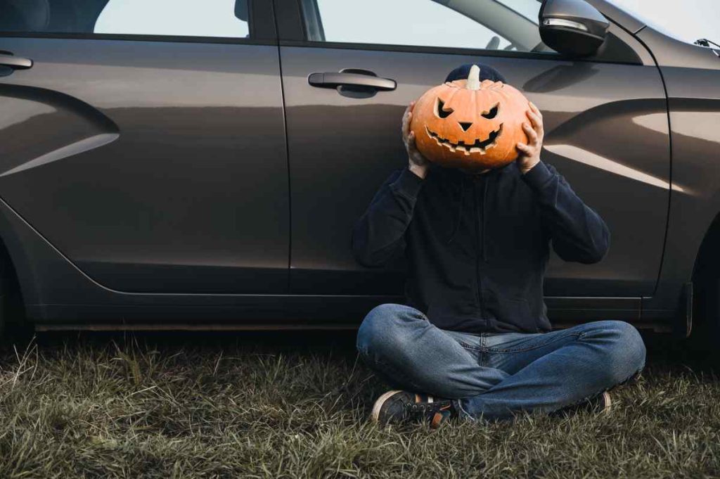 A man holds a jack-o-lantern while sitting in front of his new car