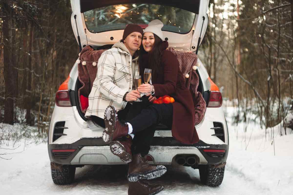 A bundled-up couple sits in the trunk of their new car in January, holding champagne flutes and snuggling for warmth