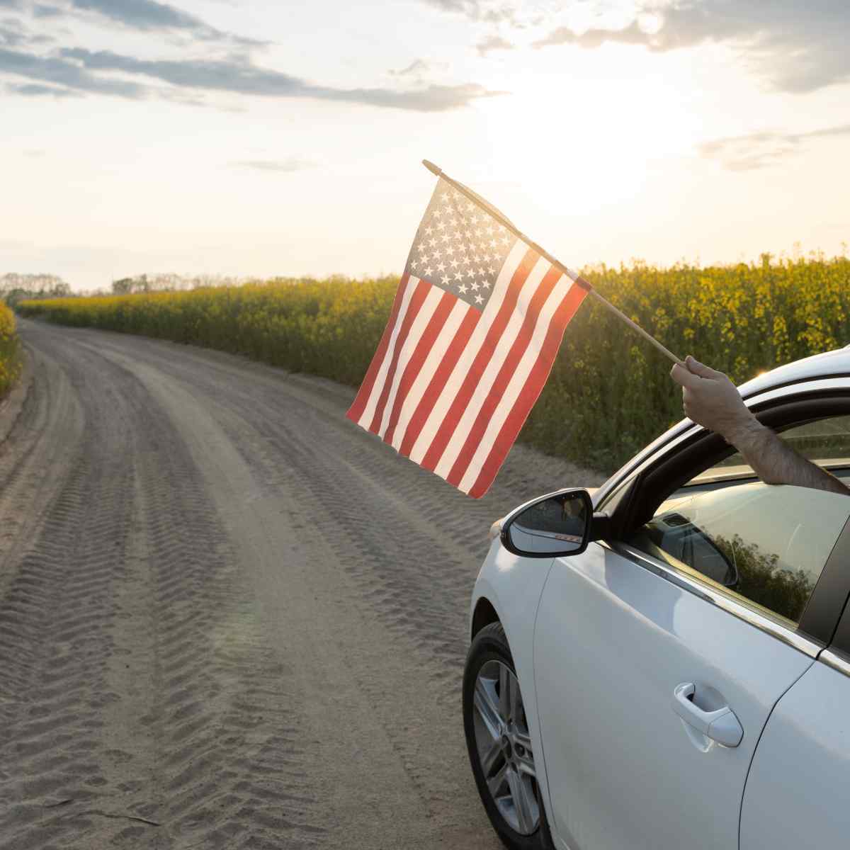 A driver holds an American flag out their car window while driving a white car on a country road.